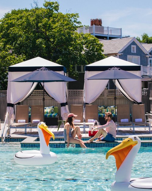 Two people relax by a pool with cabanas, surrounded by inflatable pelicans on a sunny day.