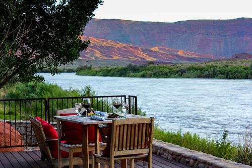 A scenic riverside dining setup with a table, chairs, and glasses, surrounded by lush greenery and mountains in the background.