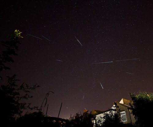 A night sky filled with stars and multiple meteor trails above a house and surrounding foliage.