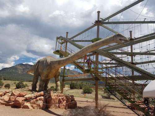 A large dinosaur sculpture with a long neck beside a climbing structure against a mountainous backdrop.