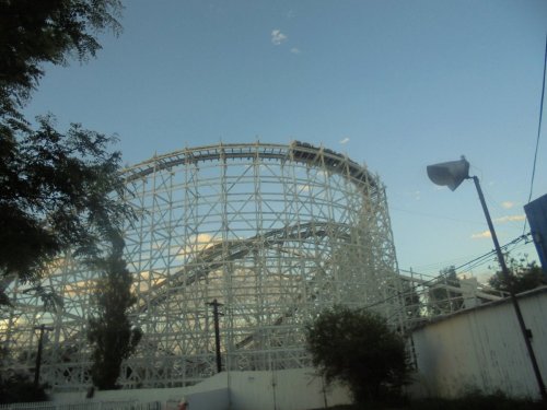 A wooden roller coaster structure against a blue sky, with trees and power lines in the foreground.
