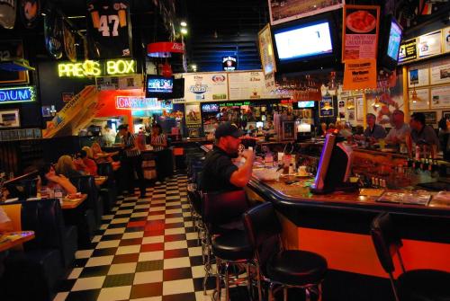 A lively sports bar with checkered floors, neon signs, and patrons enjoying food and drinks at the bar and tables.