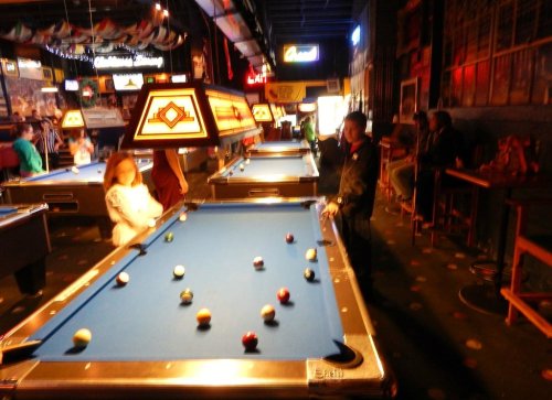 A dimly lit pool hall with several tables, players, and colorful lights creating a lively atmosphere.