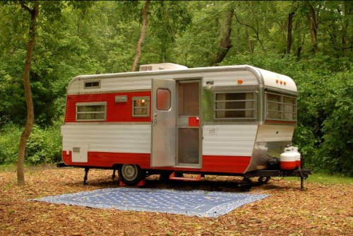 A vintage red and white camper trailer parked in a lush green forest, with a blue patterned rug laid out in front.