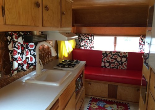 Cozy vintage kitchen with wooden cabinets, red seating, and colorful curtains, featuring a sink and stove.