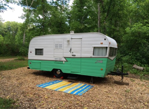 A vintage teal and white camper trailer parked on a wooded lot, with a colorful striped rug in front.
