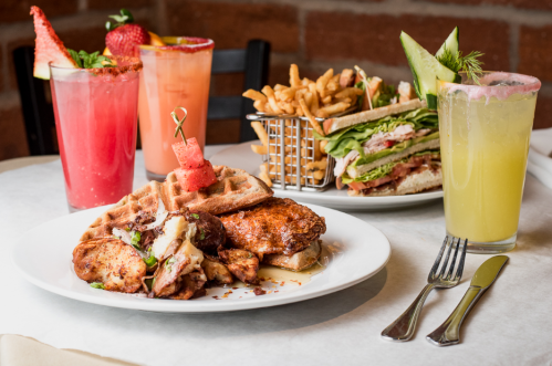 A plate with fried chicken, waffles, and potatoes, alongside drinks and a sandwich with fries in the background.