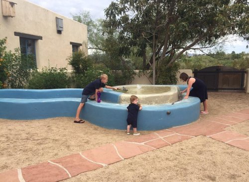 Two children and an adult interact with a small water feature in a sandy outdoor area surrounded by greenery.