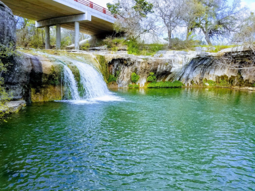 A serene waterfall cascades into a green pool beneath a bridge, surrounded by rocky terrain and sparse trees.