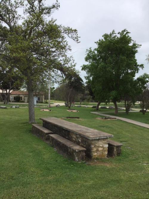 A stone picnic table surrounded by grass and trees in a park setting, with a cloudy sky above.