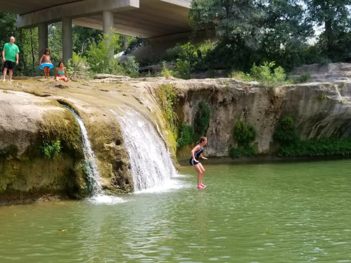 A girl prepares to jump into a green pool below a small waterfall, with people watching from the rocks above.