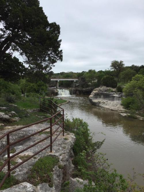 A serene river scene with a waterfall, surrounded by lush greenery and rocky formations under a cloudy sky.