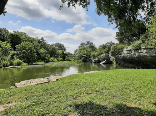 A serene riverbank scene with lush green grass, trees, and rocky outcrops under a partly cloudy sky.