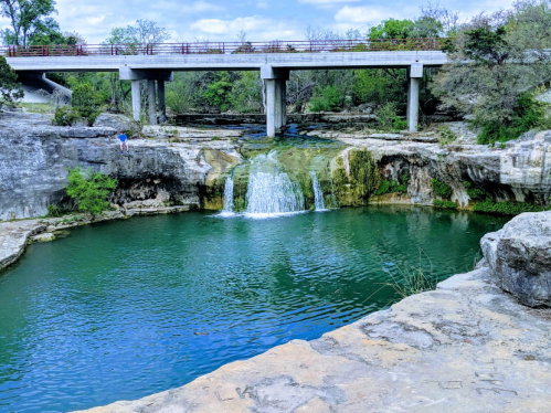 A serene waterfall cascades into a clear pool, surrounded by rocky cliffs and greenery, beneath a concrete bridge.