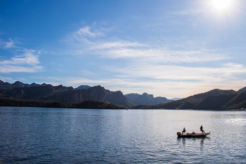 Two people fishing from a small boat on a calm lake, surrounded by mountains under a clear blue sky.