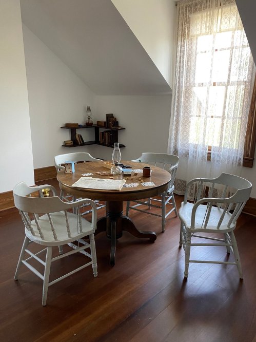 A wooden table surrounded by four light-colored chairs in a bright room with a window and shelves.