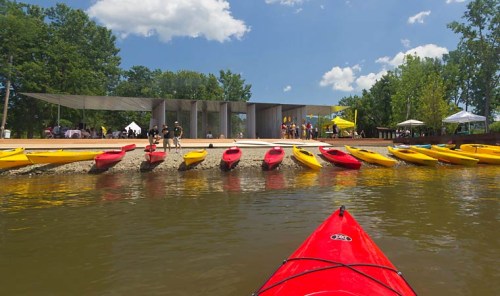 A view from a kayak towards a dock with red and yellow kayaks, surrounded by trees and a modern building in the background.