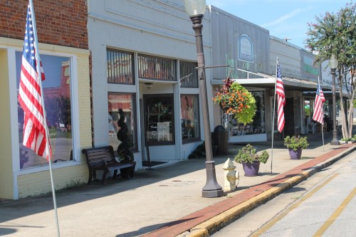 A street view of a small town with storefronts, American flags, and hanging flower baskets along the sidewalk.