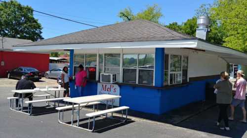 A small, colorful food stand named "Mel's" with outdoor seating and customers ordering at the window.