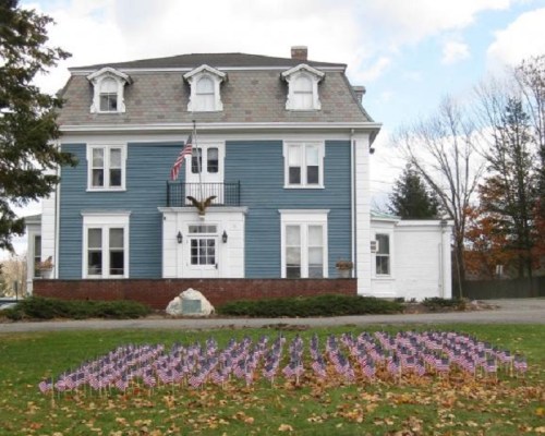 A blue house with a large eagle statue and a lawn decorated with small American flags. Trees are visible in the background.