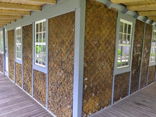 A corner of a building featuring textured walls made of natural materials, with large windows and a wooden porch.