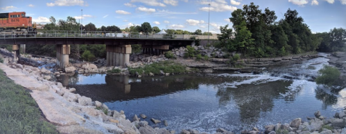 A train crosses a bridge over a river, surrounded by trees and rocky banks under a blue sky with clouds.