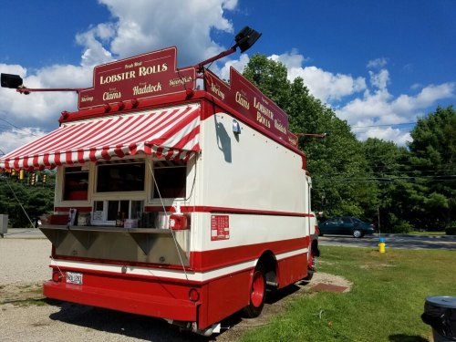 A red and white striped food truck serving lobster rolls, parked in a grassy area with trees in the background.