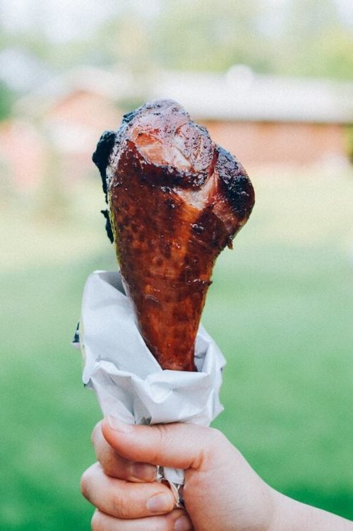 A hand holding a grilled turkey leg wrapped in paper, set against a blurred outdoor background.
