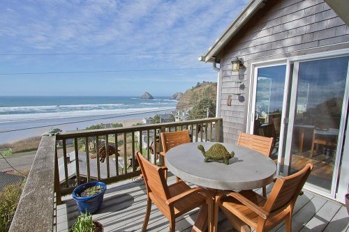 A coastal view from a deck with a round table and chairs, overlooking the ocean and distant cliffs.