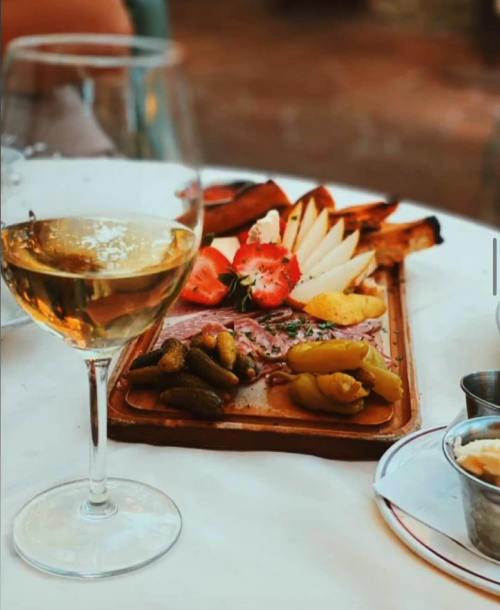 A wooden platter with assorted cheeses, meats, fruits, and pickles, alongside a glass of white wine on a table.