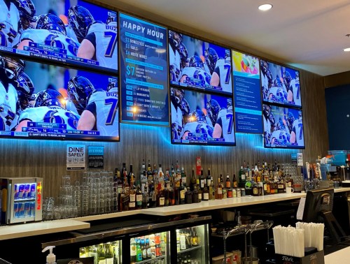 A bar with multiple screens showing a sports game, illuminated by blue lights, and a variety of drinks on the counter.