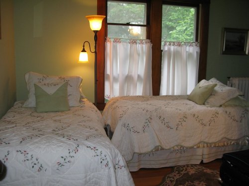 Cozy bedroom with two twin beds, floral bedding, and soft lighting from a wall lamp near a window with sheer curtains.