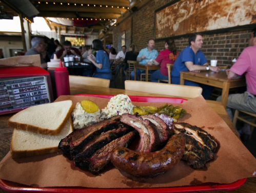 A tray of barbecue meats, sides, and bread in a bustling restaurant with diners in the background.