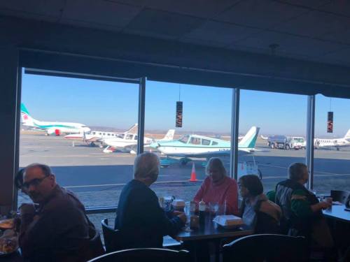A busy airport café with people dining, overlooking parked planes through large windows.