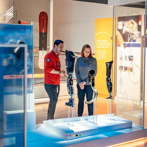 Two people examine prosthetic limbs displayed in a modern exhibit, with informative panels in the background.