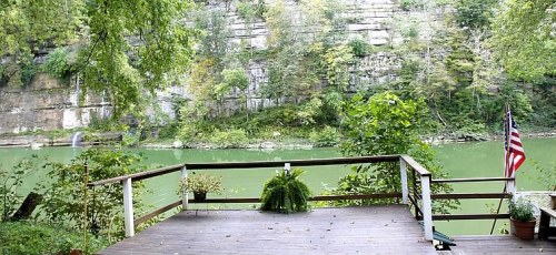 Scenic view of a river surrounded by lush greenery and cliffs, with a wooden deck and potted plants in the foreground.