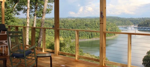 Scenic view from a wooden deck overlooking a river and lush green hills under a partly cloudy sky.