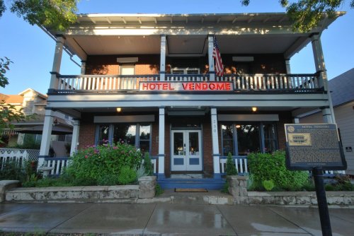 Historic Hotel Vendome with a red sign, American flag, and flower-filled garden in front. Bright blue sky above.