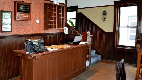 A vintage reception area featuring a wooden desk, typewriter, laptop, and staircase in a warmly lit room.