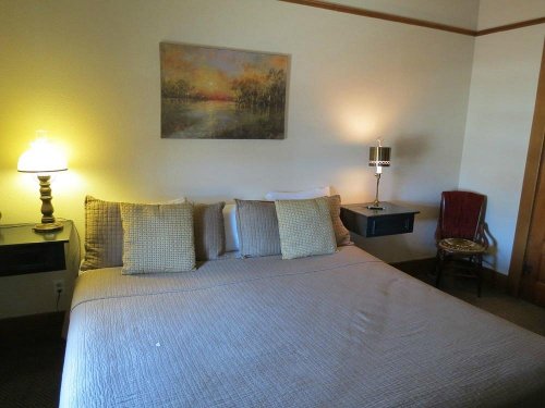Cozy bedroom with a large bed, decorative pillows, lamps, and a small chair beside a painting on the wall.