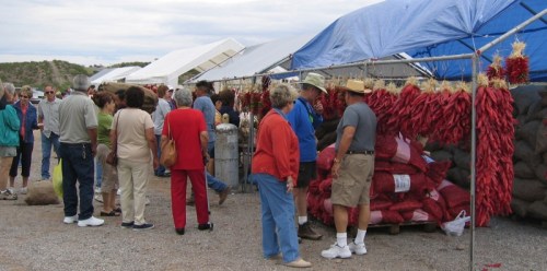 A bustling outdoor market with people browsing stalls filled with red chili peppers and other goods under blue tarps.