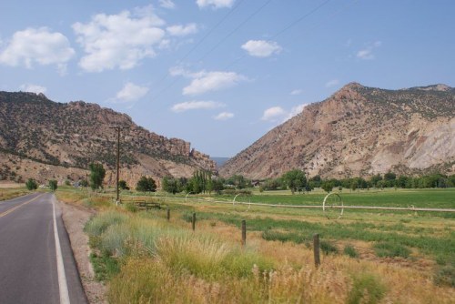 A scenic view of mountains flanking a green valley, with a road and fields in the foreground under a blue sky.