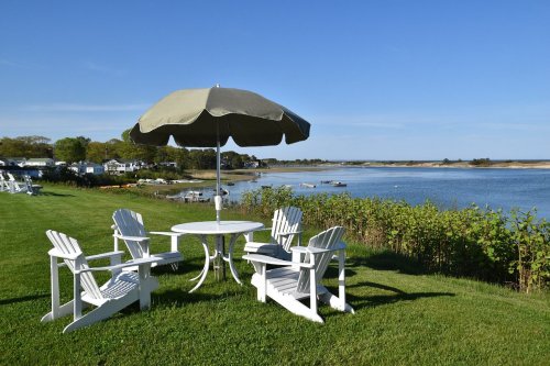 A serene lakeside scene featuring white Adirondack chairs, a table, and a large umbrella on a grassy area.