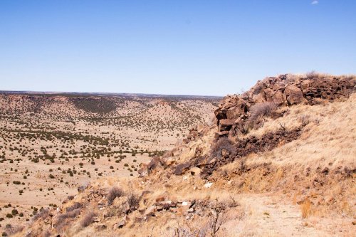 A rocky hillside overlooks a vast, arid landscape with sparse vegetation and distant hills under a clear blue sky.