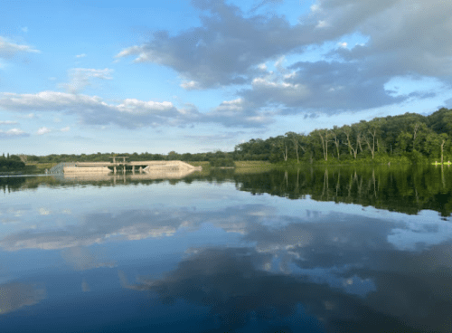 A serene lake reflecting clouds and trees, with a modern structure on the shore under a blue sky.