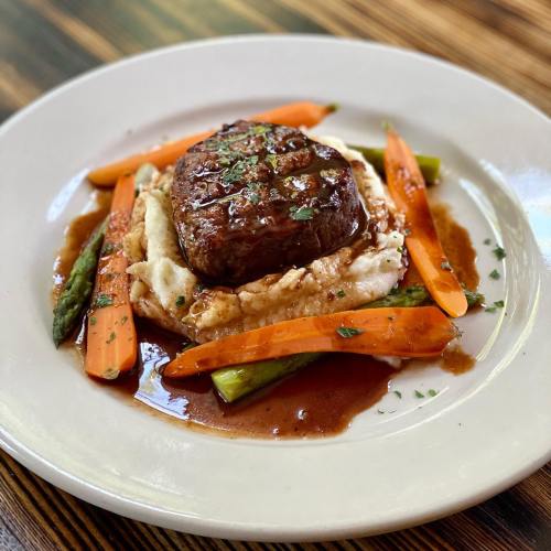 A plated dish featuring a beef filet on creamy mashed potatoes, surrounded by carrots and asparagus, drizzled with sauce.