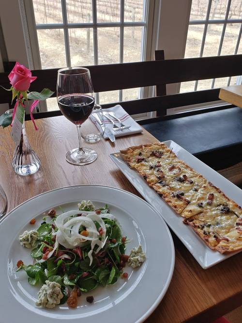 A table setting with a salad, flatbread, a glass of red wine, and a rose in a vase, with a vineyard view outside.