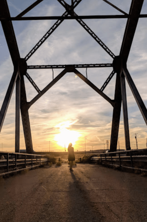 Silhouette of a person standing on a bridge at sunset, framed by the structure's metal beams.