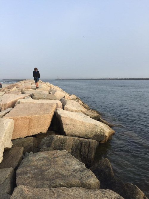 A person stands on a rocky jetty by the water, with a calm sea and hazy sky in the background.