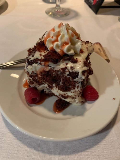 A slice of red velvet cake topped with whipped cream and drizzled with sauce, garnished with raspberries on a white plate.